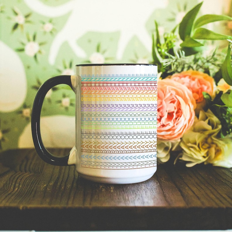 May include: White ceramic mug with a black handle and rim, decorated with a colorful, repeating pattern of stitched designs. The mug sits on a wooden surface, with flowers in the background.