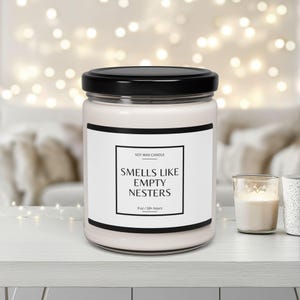 Op de afbeelding: Een witte sojawas kaars met een zwarte deksel en een label met de tekst "Soy Wax Candle" en "Smells Like Empty Nesters" met een brandtijd van 50+ uur.