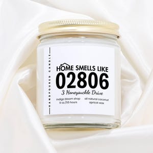 May include: A clear glass jar candle with a gold lid. The label reads "HOME SMELLS LIKE 02806 3 Honeysuckle Drive." The candle is hand-poured and made with all-natural coconut apricot wax. The candle is 9 oz. and has a burn time of 55 hours.