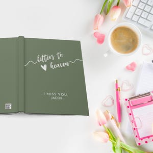 May include: A green journal with the text "letters to heaven" and a small heart. The text "I miss you, Jacob" is printed below the title. The journal is open and lying on a white surface with pink flowers, a coffee cup, and other stationery items.