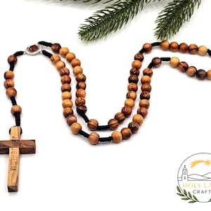 May include: A wooden rosary with olive-shaped beads and a cross pendant. The cross is engraved with the word "Jerusalem". The beads are strung on black cord. The rosary is a religious item.