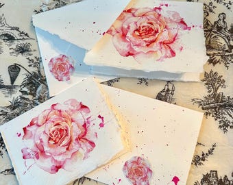 May include: A collection of handmade stationery featuring watercolor rose designs. The set includes square cards and envelopes with deckled edges, adorned with pink rose illustrations and matching rose stickers. The cards and envelopes are on a patterned background.