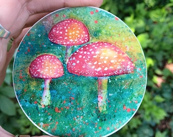 May include: A round coaster featuring a watercolor painting of three red and white spotted mushrooms. The mushrooms are set against a green and blue background with colorful paint splatters. The coaster is held in a hand.