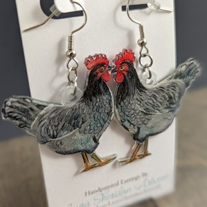 May include: A pair of handmade earrings featuring a watercolor-style illustration of two black chickens with red combs. The earrings are made of clear acrylic and have silver hooks.