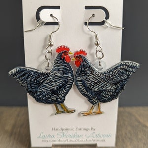 May include: A pair of hand-painted earrings featuring a detailed illustration of two roosters. The roosters have blue and white patterned feathers, red combs, and yellow beaks. The earrings hang from silver-colored hooks and are displayed on a white card.