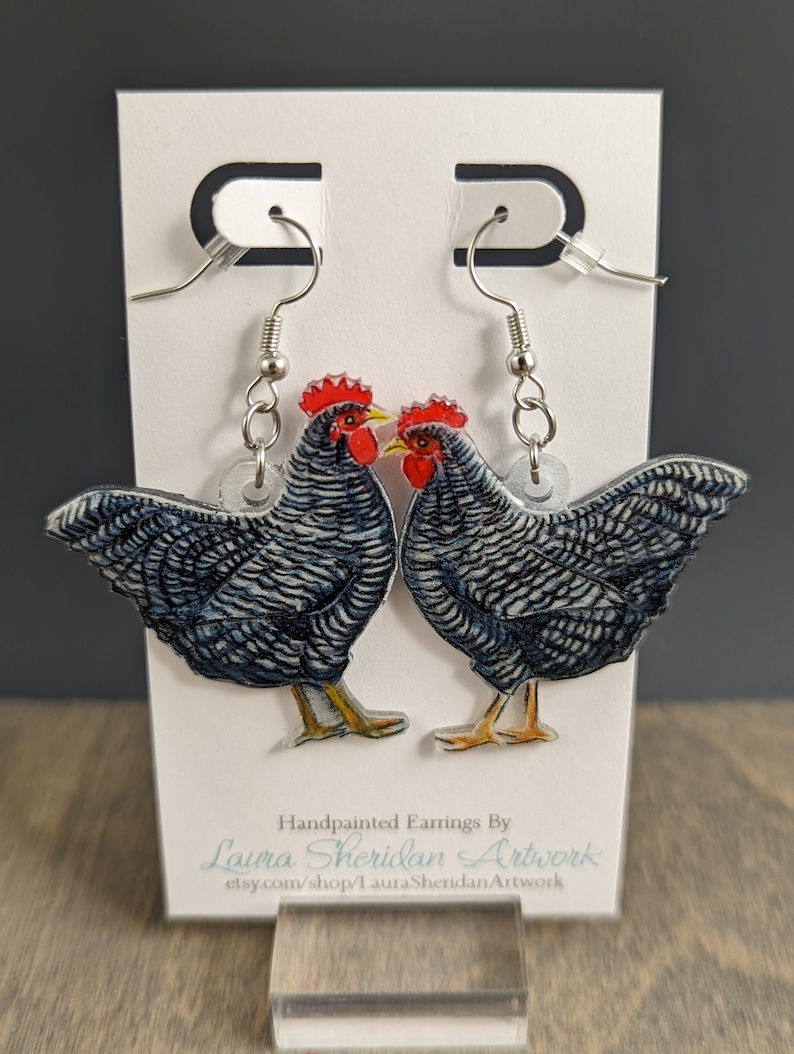 May include: A pair of hand-painted earrings featuring a detailed illustration of two roosters. The roosters have blue and white patterned feathers, red combs, and yellow beaks. The earrings hang from silver-colored hooks and are displayed on a white card.