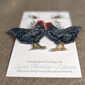 May include: Hand-painted earrings featuring two detailed chicken designs. The chickens are primarily black and white with red combs and yellow legs. The earrings are displayed on a white card with the artist's name and Etsy shop URL.