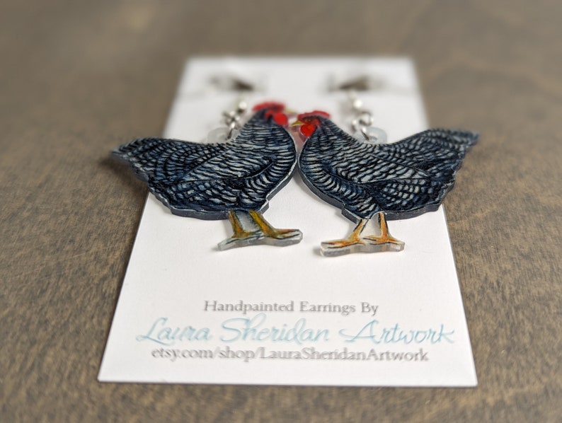 May include: Hand-painted earrings featuring two detailed chicken designs. The chickens are primarily black and white with red combs and yellow legs. The earrings are displayed on a white card with the artist's name and Etsy shop URL.