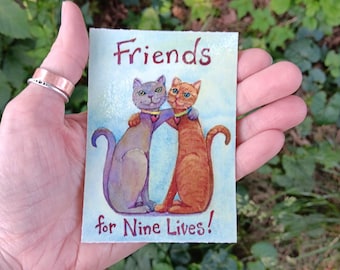 May include: A watercolor illustration of two hugging cats, one gray and one orange, with the text "Friends for Nine Lives!" in a handwritten font. The cats are wearing heart-shaped collars.