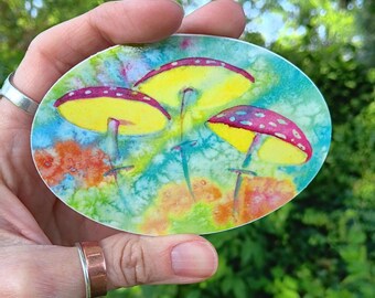 May include: Oval sticker featuring a watercolor painting of three mushrooms. The mushroom caps are yellow with red and white spots, set against a colorful background of blue, green, and orange hues. The sticker is held in a hand.