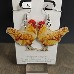 May include: A pair of hand-painted earrings featuring two chickens facing each other. The chickens are yellow and brown with red combs and waddles. The earrings hang from silver-colored hooks. The text "Handpainted Earrings By Laura Sheridan Artwork" is visible.