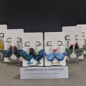 May include: A collection of acrylic earrings featuring colorful rooster and hen designs. Each pair of earrings is displayed on a white card with a silver hook. The text "ChickieRINGS & COCKRINGS" and "Create Your Own Flock" is visible.