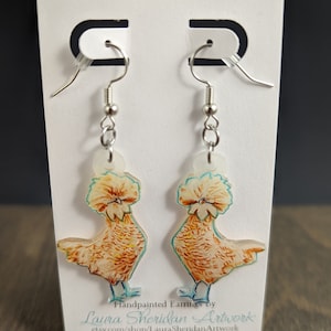 May include: Handpainted earrings featuring a pair of chickens with fluffy white crests and brown bodies. The earrings are made of clear acrylic and have silver hooks. The earrings are displayed on a white card with the text "Handpainted Earrings by Laura Sheridan Artwork etsy.com/shop/LauraSheridanArtwork".