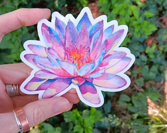 May include: A colorful sticker featuring a watercolor illustration of a lotus flower. The flower has shades of pink, purple, and blue, with a white border. The sticker is held against a backdrop of green foliage.