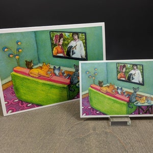 May include: Two art prints depict a cozy scene of cats watching TV. The cats lounge on a green couch with a pink cushion, in a room with a teal wall and a patterned rug. A lamp with blue and gold globes stands nearby. The TV shows a couple in period clothing.