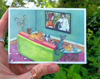 May include: A colorful illustration depicts cats watching TV. The cats are lounging on a green couch with a pink and purple rug. The TV shows two people. A small table has snacks. The artwork is held by a hand.