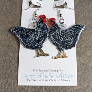 May include: Hand-painted earrings featuring two chickens with black and white striped feathers, red combs, and yellow feet. The earrings hang from silver-colored hooks and are displayed on a white card with the artist's name and Etsy shop URL.