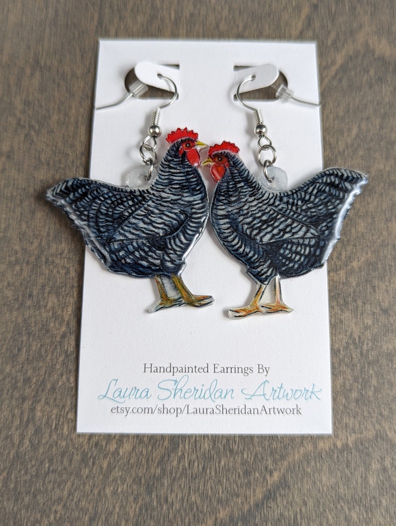 May include: Hand-painted earrings featuring two chickens with black and white striped feathers, red combs, and yellow feet. The earrings hang from silver-colored hooks and are displayed on a white card with the artist's name and Etsy shop URL.