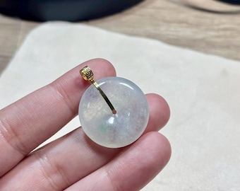 May include: A round, pale green jade pendant with a gold-colored bail. The jade has a translucent quality, with subtle variations in color. The pendant is held by a hand, showcasing its size and design. The bail has a decorative pattern.