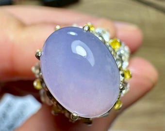 May include: Oval-shaped lavender gemstone ring with a silver band. The gemstone has a gradient of colors, transitioning from a light purple to a soft pink. The ring is adorned with small yellow and clear stones around the main gem.