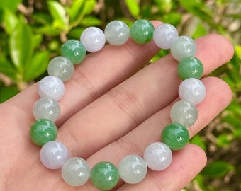May include: A beaded bracelet featuring alternating green and white jade spheres. The bracelet is held in a hand, with a blurred green foliage background. The jade beads are polished and have a smooth, reflective surface. The bracelet is a piece of jewelry.