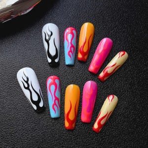 May include: Ten long, coffin-shaped artificial nails in various colours. Each nail features a flame design in contrasting colours. The colour schemes include white with black flames, blue with pink flames, orange with red flames, and pink with orange flames.