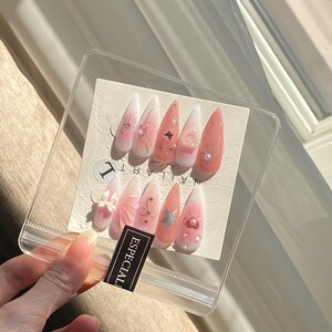 May include: A set of ten long, almond-shaped artificial nails in a clear plastic package. The nails feature a gradient design with pink and white hues, adorned with various embellishments like silver glitter, pearls, and floral accents. The word "ESPECIAL" is printed on a black label.