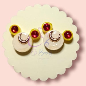 Puede incluir: Un par de pendientes con una luna creciente blanca con un dije de luna creciente dorado y un círculo amarillo con una gema roja en el centro.