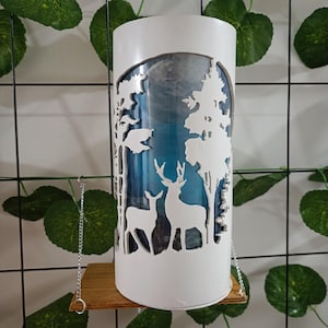 Puede incluir: Portavelas cilíndrico blanco con un diseño recortado de dos ciervos y árboles. El portavelas tiene un fondo azul y una luna blanca.