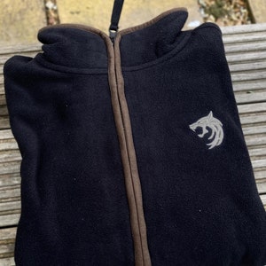 May include: Black fleece jacket with a full zipper and a high collar. The jacket has a brown trim along the zipper and collar. A silver wolf head emblem is embroidered on the left chest. The jacket is folded and lying on a wooden surface.