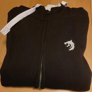 May include: Black zippered hoodie with a white wolf logo embroidered on the front. The hoodie has a white drawstring and a white lining around the hood.