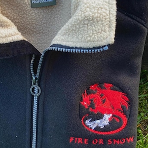 May include: Black fleece jacket with a cream-colored interior lining and a full zipper. The jacket features a red dragon and white wolf emblem with the text "FIRE OR SNOW" embroidered on the front. The jacket has the "REGATTA PROFESSIONAL" logo.