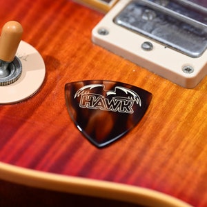 Puede incluir: Primer plano de una púa de guitarra marrón y negra con la palabra "HAWK" impresa en blanco.