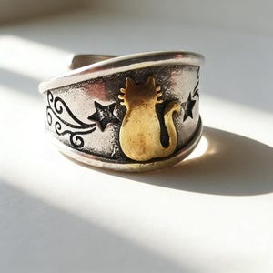 Puede incluir: Anillo plateado con un diseño de gato y estrellas doradas. El anillo presenta un patrón grabado y un acabado texturizado. El gato está sentado y las estrellas están dispersas alrededor del gato. Es una pieza de joyería.
