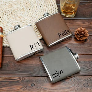 Puede incluir: Tres petacas personalizadas en cuero beige, marrón y gris, cada una con ribete plateado. Las petacas están grabadas con iniciales y un nombre. También se ven un cigarro, una piña y un vaso de líquido ámbar.