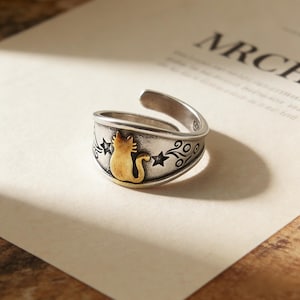 May include: A silver-toned adjustable ring featuring a gold-colored cat silhouette. The ring is decorated with star and swirl designs. The cat is centered on the ring's face, with the adjustable band curving upwards.