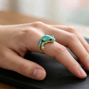 Puede incluir: Un anillo con forma de pequeña rana con un acabado liso en color azul verdoso claro. La rana está colocada de modo que su cuerpo envuelve el dedo, con las patas delanteras y traseras extendiéndose a lo largo de la banda. Tiene ojos oscuros.