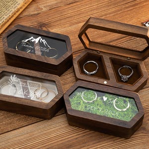 Könnte beinhalten: Holzringboxen mit transparenten Deckeln, die Ringe und personalisierte Texte zeigen. Eine Box trägt die Aufschrift "Tammi & Tiller" und "Sep 5, 2025". Eine andere Box hat "J & K" und "Aug 7, 2025". Eine dritte Box hat "Maleale Jackson" und "Sep 7, 2025".