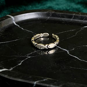 May include: A silver-colored adjustable ring featuring a fish design. The ring has an open design and is decorated with multiple fish motifs around the band. The ring is displayed on a black marble surface.