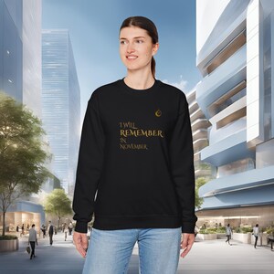 May include: Black sweatshirt with gold text reading "I Will Remember in November". A small gold emblem is on the upper right chest. The person is wearing blue jeans. The background shows a modern cityscape.