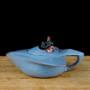 Könnte beinhalten: Eine blaue Teekanne aus Keramik mit einem einzigartigen Design, auf der zwei kleine Figuren sitzen. Die Teekanne hat einen Ausguss und einen Griff, und die Oberfläche ist mit kleinen Punkten strukturiert.