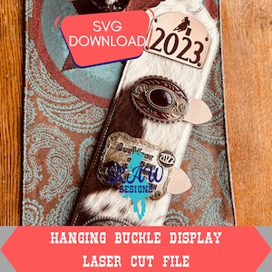 May include: A white and brown cowhide hanging display with two silver belt buckles. The top buckle has a red stone and the bottom buckle says "Out West 2022" and "Raw Designs". A white tag with the number "2023" is attached to the display.