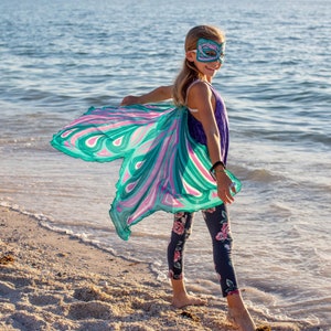 May include: A young person wearing a purple tank top and floral leggings with a teal and pink butterfly wings costume and a teal butterfly mask. The person is standing on a sandy beach with the ocean in the background.