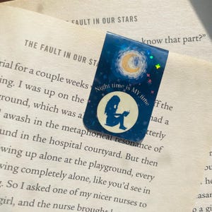 以下が含まれることがあります： 三日月と星が描かれた青と銀のしおり。しおりには、本を読んでいる人のシルエットと「Night time is MY time」という文字が書かれています。