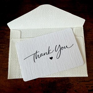 May include: A white textured envelope with a matching card. The card features the words "Thank You" in elegant script with a small heart. The envelope is open, revealing the card inside, set against a dark wood background.