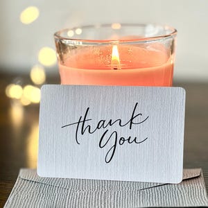 May include: A burning peach-coloured candle in a clear glass jar is behind a white card and envelope. The card says "thank You" in elegant black script. The background has soft, out-of-focus lights.