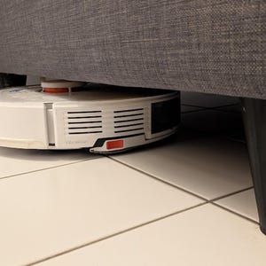Könnte beinhalten: Ein weiß-schwarzer Roboterstaubsauger mit orangefarbenem Akzent befindet sich unter einem grauen Sofa. Der Staubsauger steht auf einem weiß gefliesten Boden. Der Staubsauger trägt den Text "VibraRise".
