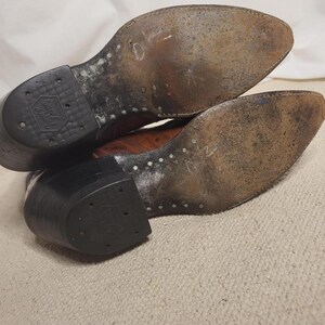 May include: Pair of well-worn brown leather cowboy boots, soles facing up. The soles show signs of wear and use, with visible stitching and a rubber heel. The boots have a classic Western design.