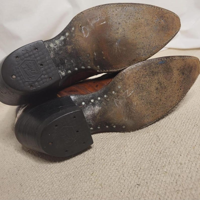 May include: Pair of well-worn brown leather cowboy boots, soles facing up. The soles show signs of wear and use, with visible stitching and a rubber heel. The boots have a classic Western design.