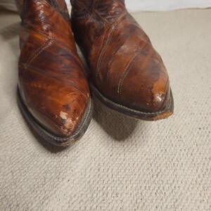 May include: A pair of well-worn brown leather cowboy boots. The boots have a pointed toe and a stitched design. The leather shows signs of wear and tear, with some scuffing and discoloration. The boots are set against a neutral background.